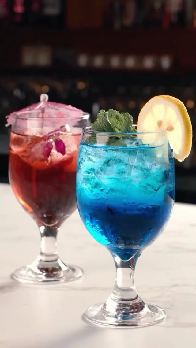 Two cocktails on the marble bar, one blue with lemon and mint, one red with a paper parasol